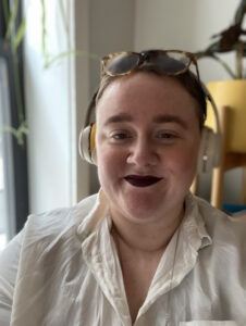 Selfie of Jesse Rice-Evans, a fat white femme, wearing gold over-ear headphones, a white button-down, and glasses pushed up on her head. She wears a dark red lip color and smiles without showing her teeth.