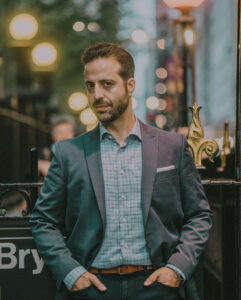 Haralabos [Harry] Stafylakis wears a gray blazer over a blue collared dress shirt, hands in pockets, head turned to the side, looking directly in the camera, in front of a subway station with city lights blurry in the background